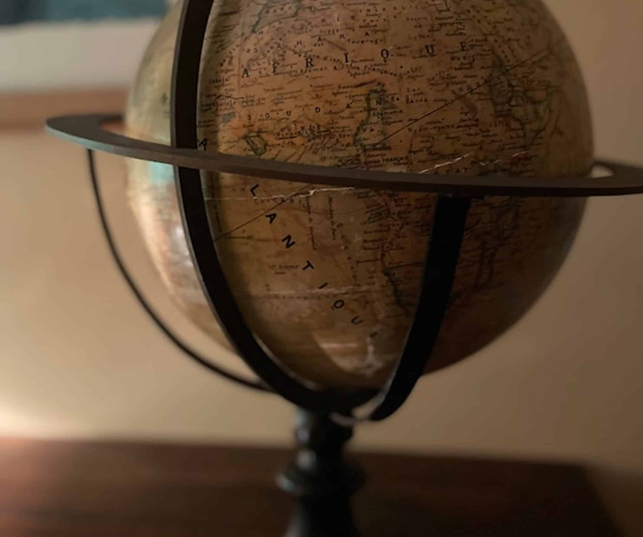Un globe terrestre vieilli montre les continents, avec "AFRIQUE" et "L'ATLANTIQUE" visibles. Il est posé sur un support sombre, dans une lumière tamisée.