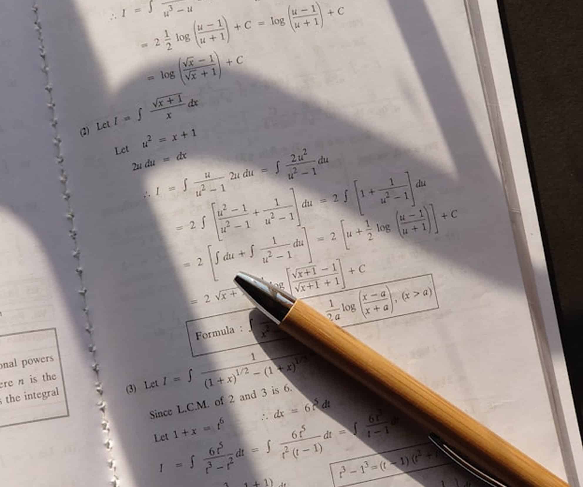 Un stylo couleur bambou repose en diagonale sur un manuel ouvert à spirale, affichant des équations et formules de calcul intégral sous une forte ombre diagonale. Texte sur l'image : = ½ log (u - 1 / u + 1) + C = log (u - 1 / u + 1) = log (√x - 1 / √x + 1) + C (2) Let I = ∫ √x + 1 / x dx Let u² = x + 1 2u du = dx ∴ I = ∫ u / (u² - 1) 2u du = ∫ 2u² / (u² - 1) du = 2 ∫ [ (u² - 1 + 1) / (u² - 1) ] du = 2 ∫ [ 1 + 1 / (u² - 1) ] du = 2 [ ∫ du + ∫ 1 / (u² - 1) du ] = 2 [ u + ½ log (u - 1 / u + 1) ] + C Formula: 1 / (2a) log (x - a / x + a), (x > a) onal powers re n is the s the integral (3) Let I = ∫ 1 / [ (1 + x)¹/² - (1 + x)¹/³ ] Since L.C.M. of 2 and 3 is 6. Let 1 + x = t⁶ ∴ dx = 6t⁵ dt I = ∫ 6t⁵ / (t³ - t²) dt = ∫ 6t⁵ / [ t² (t - 1) ] dt t³ - 1³ = (t - 1) (t² + t + 1)