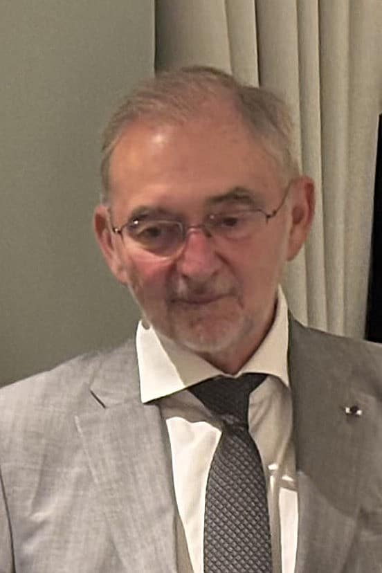 Un homme d'âge mûr aux cheveux gris et lunettes, en costume gris, chemise blanche et cravate à motifs, regarde vers le bas. Le fond est clair et uni.