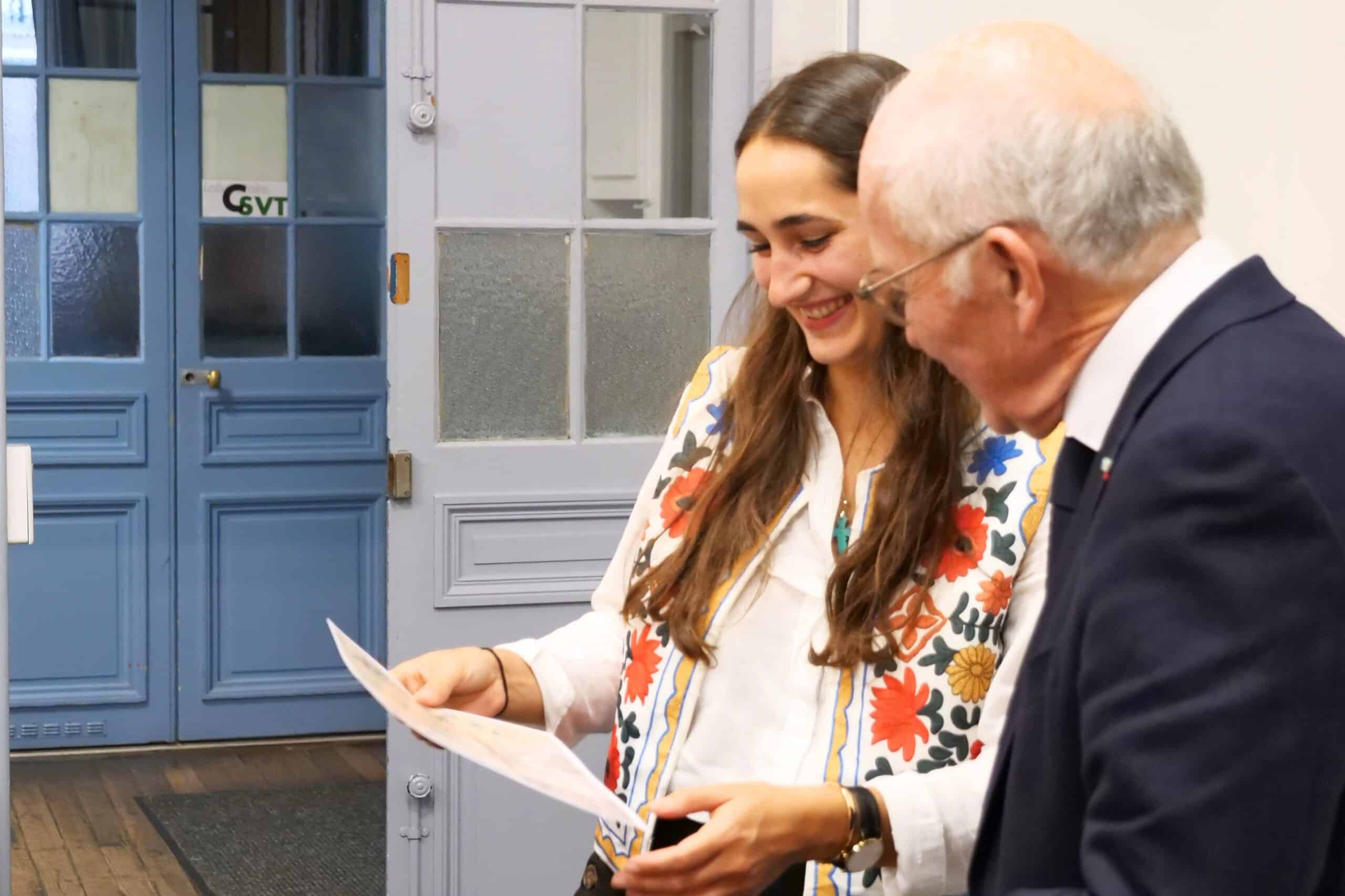 Une jeune femme et un homme âgé regardent en souriant un document, debout devant des portes bleues intérieures, dont une affiche : C6VT.