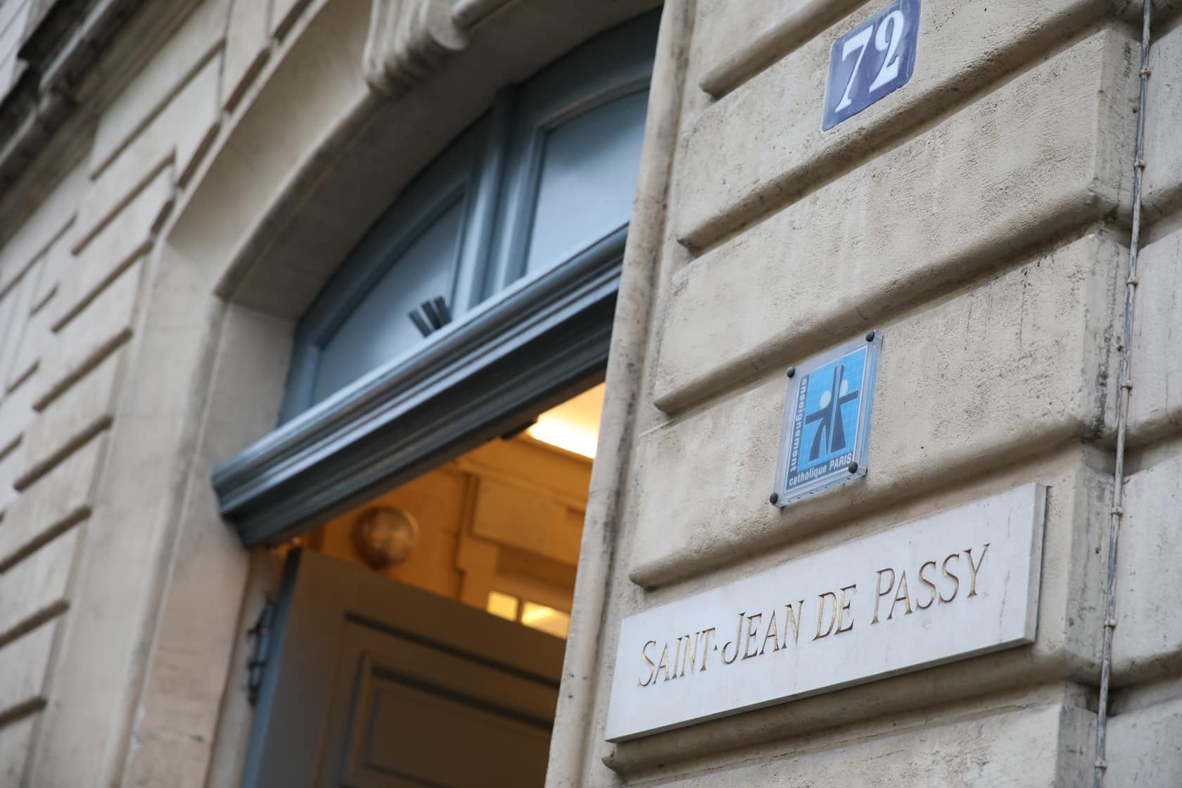 Entrée de bâtiment en pierre ornée avec une porte grise semi-ouverte. L'intérieur est éclairé. Des plaques affichent "72", un logo "enseignement catholique PARIS" et "SAINT-JEAN DE PASSY".
