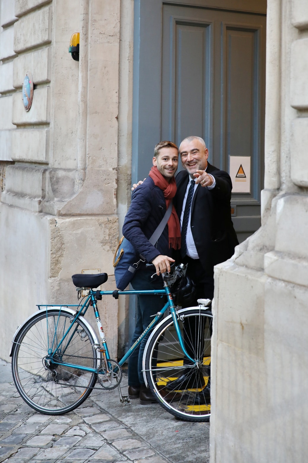 Deux hommes souriants posent devant une porte grise. L'aîné, en costume, enlace le plus jeune qui tient un vélo turquoise PEUGEOT et pointe l'observateur. Contexte urbain en pierre.