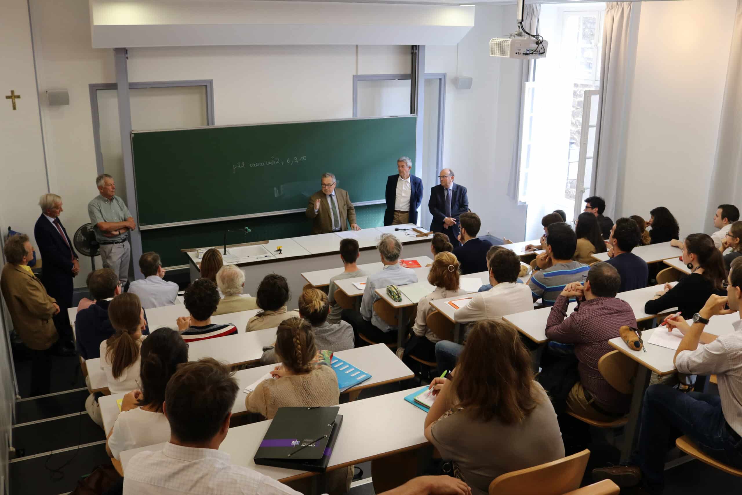 Un orateur s'adresse à une trentaine d'étudiants et quelques adultes dans un amphithéâtre moderne. Au tableau vert: "p22 exercices 2, 6, 9, 10". Un crucifix est au mur. Sur un classeur, on lit "NOMAD".