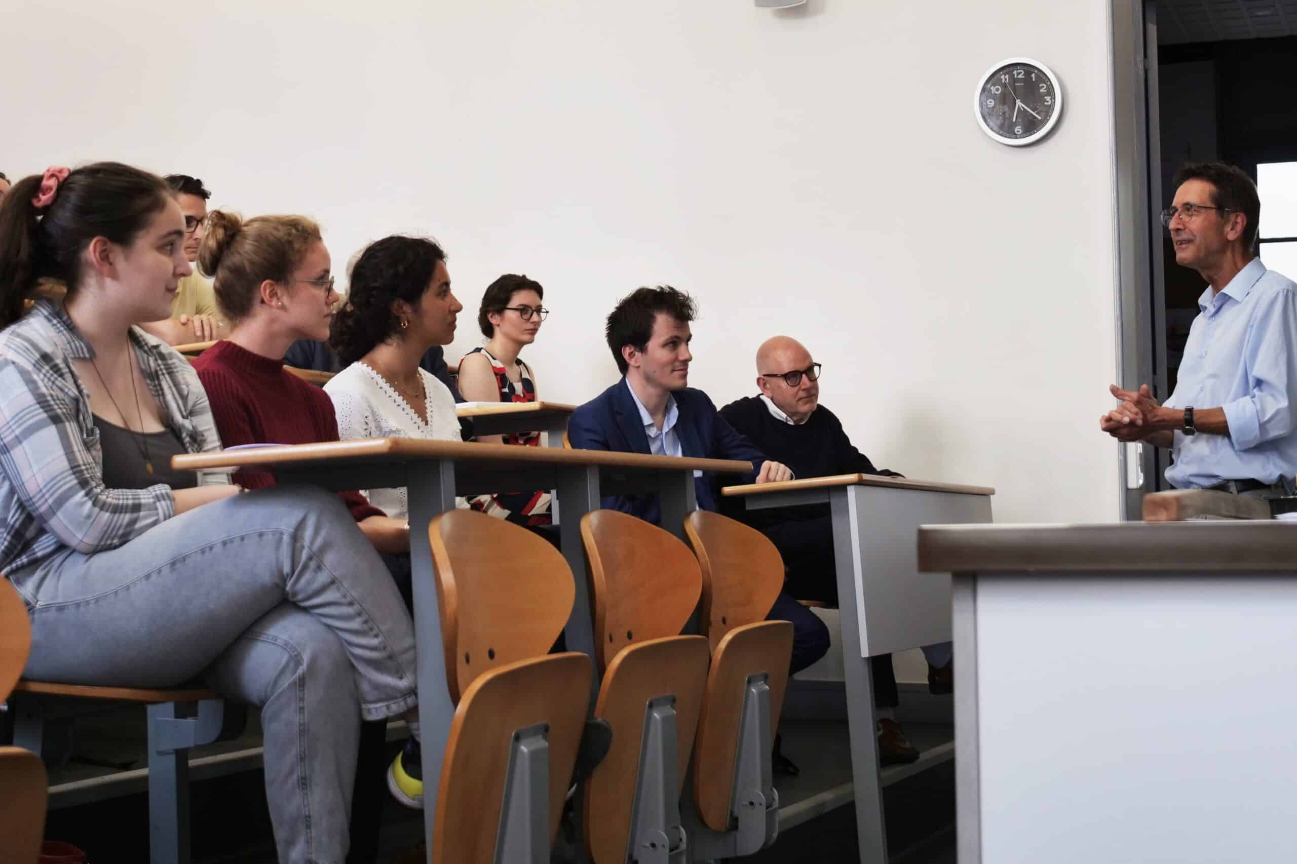 Un professeur parle à un groupe de personnes assises en rangées dans une salle de cours. Une horloge murale noire et blanche, affichant environ 11h45, est visible à droite.