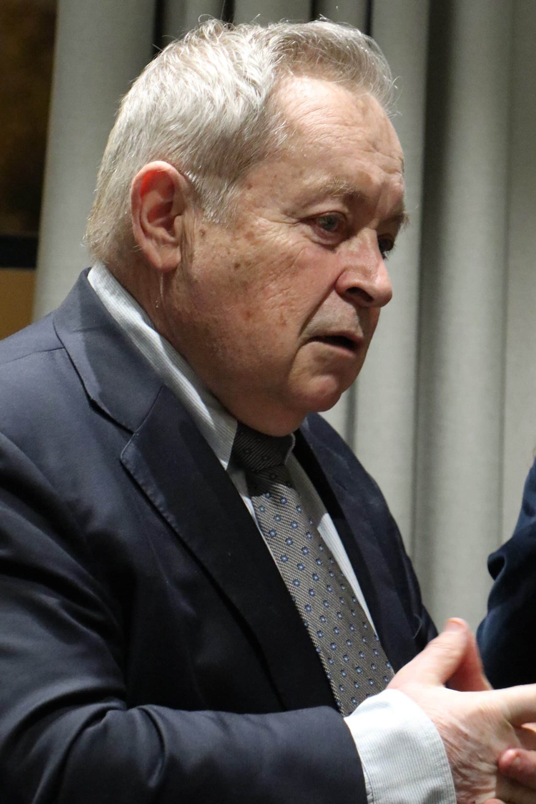 Un homme âgé aux cheveux gris, vêtu d'un costume sombre et d'une cravate à motifs, regarde vers le bas, les mains jointes, devant un arrière-plan gris flou.