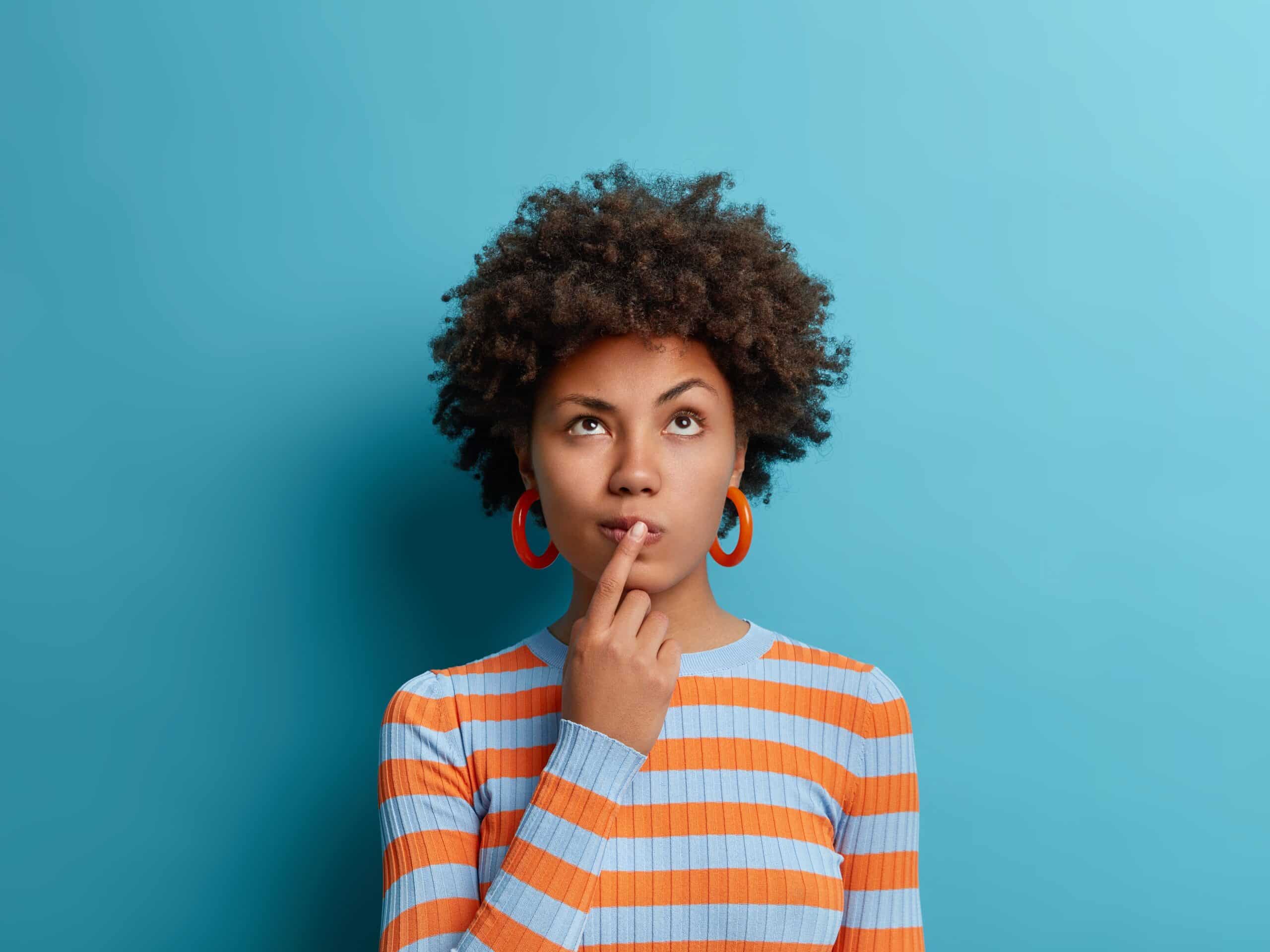 Une jeune femme aux cheveux bouclés foncés réfléchit, le doigt sur le menton, regardant vers le haut. Elle porte un pull rayé bleu et orange et des créoles, sur fond bleu uni.
