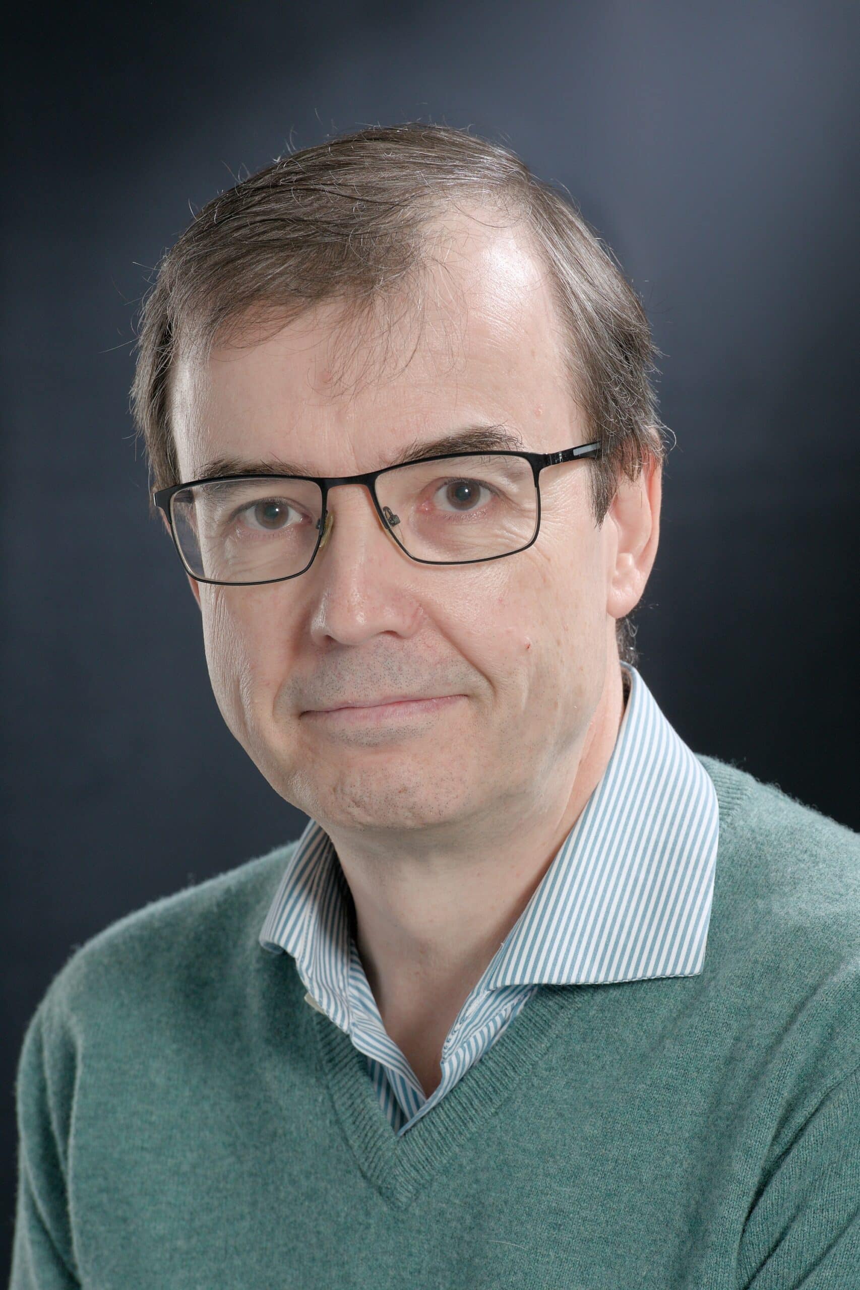 Un homme d'âge moyen, portant des lunettes et un pull vert d'eau sur une chemise rayée, sourit légèrement en regardant vers l'avant sur un fond gris uni.
