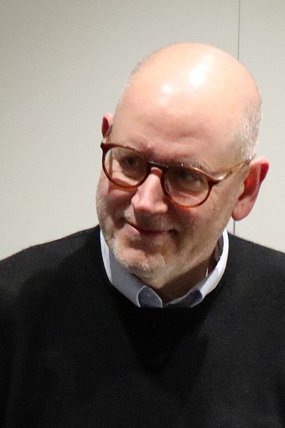 Homme chauve d'âge moyen portant lunettes et barbe légère, regardant de côté avec un sourire discret. Il porte un pull noir sur une chemise claire devant un fond uni.