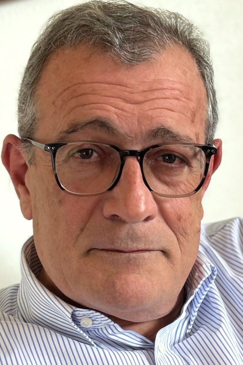 Un homme aux cheveux gris et lunettes regarde frontalement. Il porte une chemise rayée bleue et blanche, son visage est cadré serré sur un fond clair.